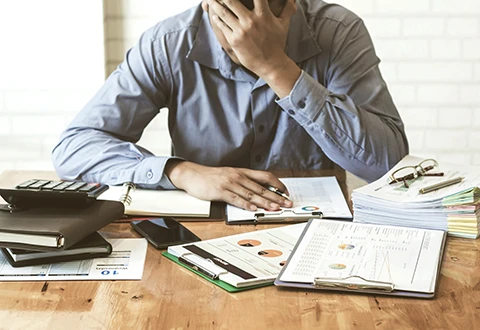 Homem com expressão de stress rodeado de relatórios, papéis e calculadora. Representa a pressão de lidar com avaliações negativas e evitar penalizações na Amazon segundo as novas regras.