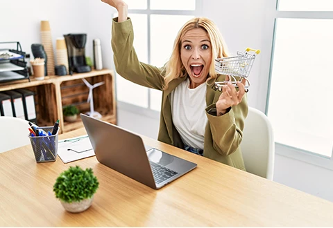 Imagem de uma mulher loira, sorridente, a segurar um mini carrinho de compras enquanto trabalha no portátil. Representa sucesso em e-commerce, marketing digital ou gestão de loja online.